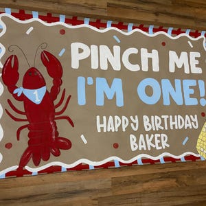 May include: A rectangular sign with a brown background, red and white border, and a red crawfish illustration. Text reads "Pinch Me I'm One! Happy Birthday Baker."