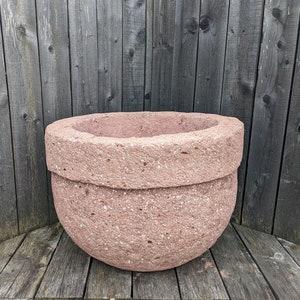 May include: A large, round, pink planter made of rough textured stone. The planter is sitting on a wooden surface.