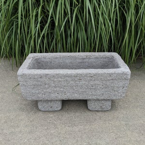 May include: A gray concrete planter with a rectangular shape and four short legs. The planter has a rough, textured surface.