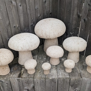 Pu&ograve; includere: Un set di otto sgabelli da giardino in cemento a forma di fungo, di diverse dimensioni, che vanno dal piccolo al grande. Gli sgabelli sono tutti di colore grigio chiaro e hanno una superficie ruvida e testurizzata.