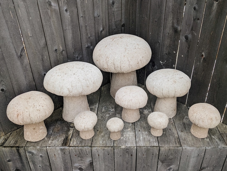 Pu&ograve; includere: Un set di otto sgabelli da giardino in cemento a forma di fungo, di diverse dimensioni, che vanno dal piccolo al grande. Gli sgabelli sono tutti di colore grigio chiaro e hanno una superficie ruvida e testurizzata.