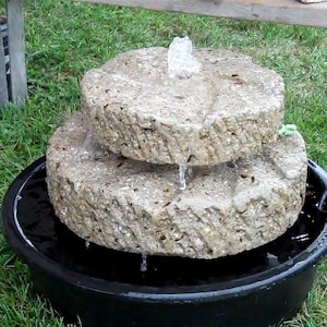 May include: A two-tiered stone fountain with a black basin, set on a grassy lawn. Water flows from the top tier, creating a cascading effect.
