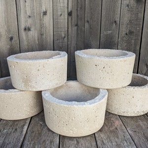 May include: Five round concrete planters in various sizes, stacked on a wooden surface. The planters are a light beige color and have a rough, textured surface.