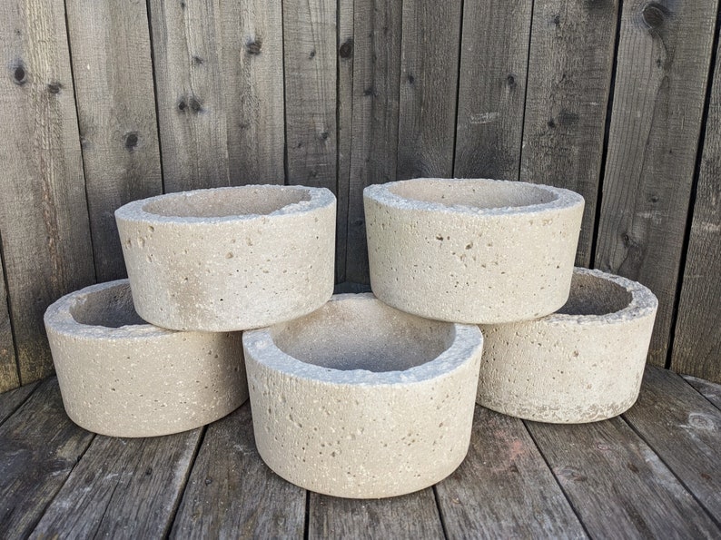 May include: Five round concrete planters in various sizes, stacked on a wooden surface. The planters are a light beige color and have a rough, textured surface.