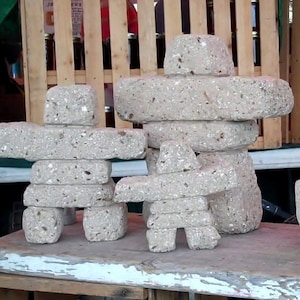 May include: Two stone sculptures of Inukshuk figures, one larger than the other, are displayed on a wooden table. The sculptures are made of stacked stones and have a rough, textured surface.