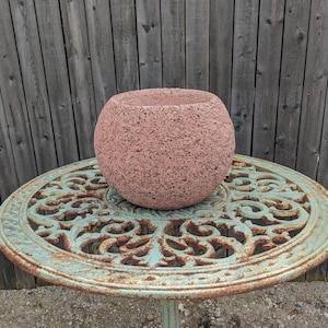 May include: A round, terracotta-colored planter with a rough, textured surface sits on a green metal table with a decorative, ornate design. The table has a circular top and a single leg.