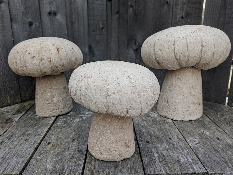 Pu&ograve; includere: Tre sgabelli da giardino in cemento a forma di fungo di diverse dimensioni. Gli sgabelli sono di colore beige chiaro e hanno una superficie testurizzata.