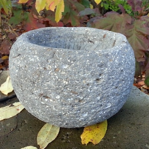 May include: A round, gray concrete planter with a rough, textured surface. The planter is empty and sits on a gray concrete surface.