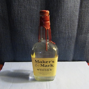May include: A clear glass Maker's Mark whisky bottle with a cream-colored label featuring black text and a distinctive red wax seal dripping down the neck of the bottle. The bottle is partially filled with a light amber liquid and has a square base.