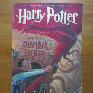 May include: A hardcover book titled "Harry Potter and the Chamber of Secrets" by J.K. Rowling. The cover features a red dragon-like creature and a boy with glasses. The book is a Scholastic edition.