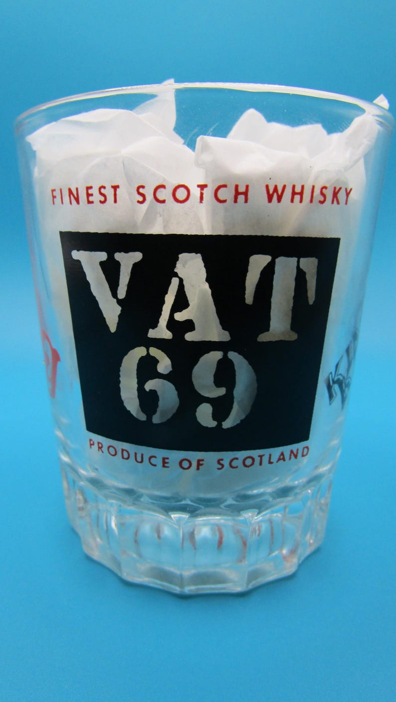 Może przedstawiać: Przezroczysty szklany kubek z napisem "FINEST SCOTCH WHISKY" i "VAT 69 PRODUCE OF SCOTLAND". Szklanka jest wypełniona białym papierem i stoi na niebieskim tle.