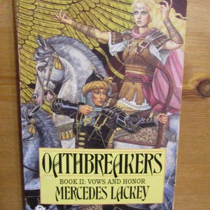 Peut inclure: Couverture du livre Oathbreakers, Livre II : Vœux et Honneur de Mercedes Lackey. La couverture représente une femme en armure chevauchant un cheval blanc sur un fond d'ailes dorées. La femme tient un arc et une flèche. Le titre du livre est en grandes lettres grasses.
