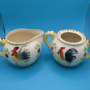 May include: Creamer and sugar bowl set. The set features a rooster design with red, orange, and black details, along with green leaves and purple berries. The handles are yellow with brown stripes. The interior of the creamer and sugar bowl is beige.