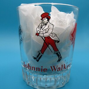 Może przedstawiać: Przezroczysty szklany kubek z logo Johnnie Walker. Logo przedstawia mężczyznę w czerwonym płaszczu i kapeluszu, idącego z laską. Na szkle widnieje napis "Johnnie Walker".