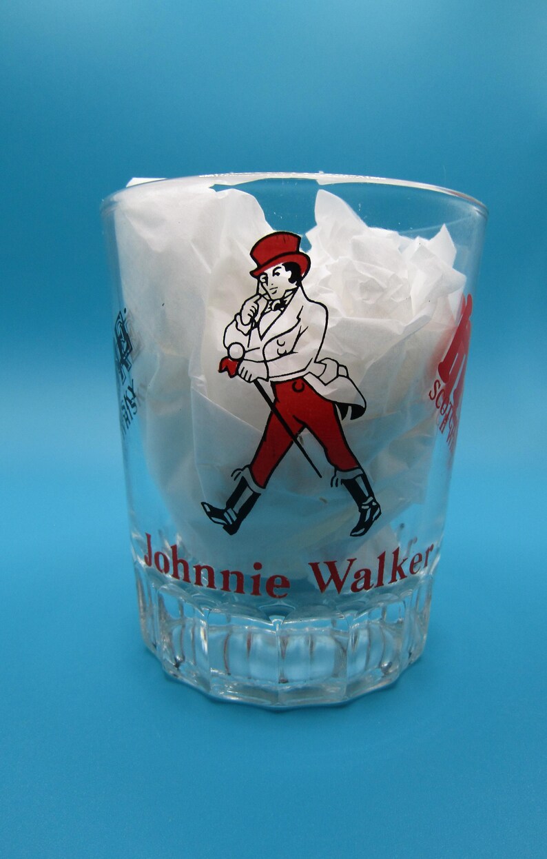 Może przedstawiać: Przezroczysty szklany kubek z logo Johnnie Walker. Logo przedstawia mężczyznę w czerwonym płaszczu i kapeluszu, idącego z laską. Na szkle widnieje napis "Johnnie Walker".