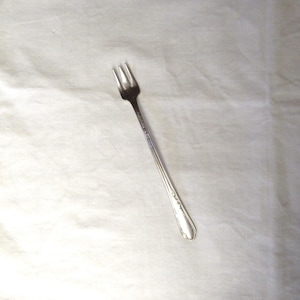 May include: A long, slender silver-toned cocktail fork featuring three tines and a smooth, tapered handle with a subtle decorative embossed pattern near the base. The metal surface has a polished, reflective finish throughout the entire utensil.