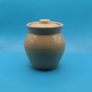 May include: A small, beige ceramic jar with a matching lid. The jar has a rounded body and a slightly flared rim. The lid has a small knob on top for easy removal. The jar is set against a solid blue background.