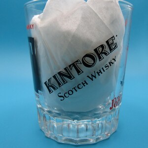 Może przedstawiać: Przezroczysty kieliszek do w&oacute;dki z czarnym napisem "Kintore Scotch Whisky". Kieliszek jest wypełniony białą bibułą i stoi na niebieskim tle. Kieliszek ma wzorzystą podstawę.