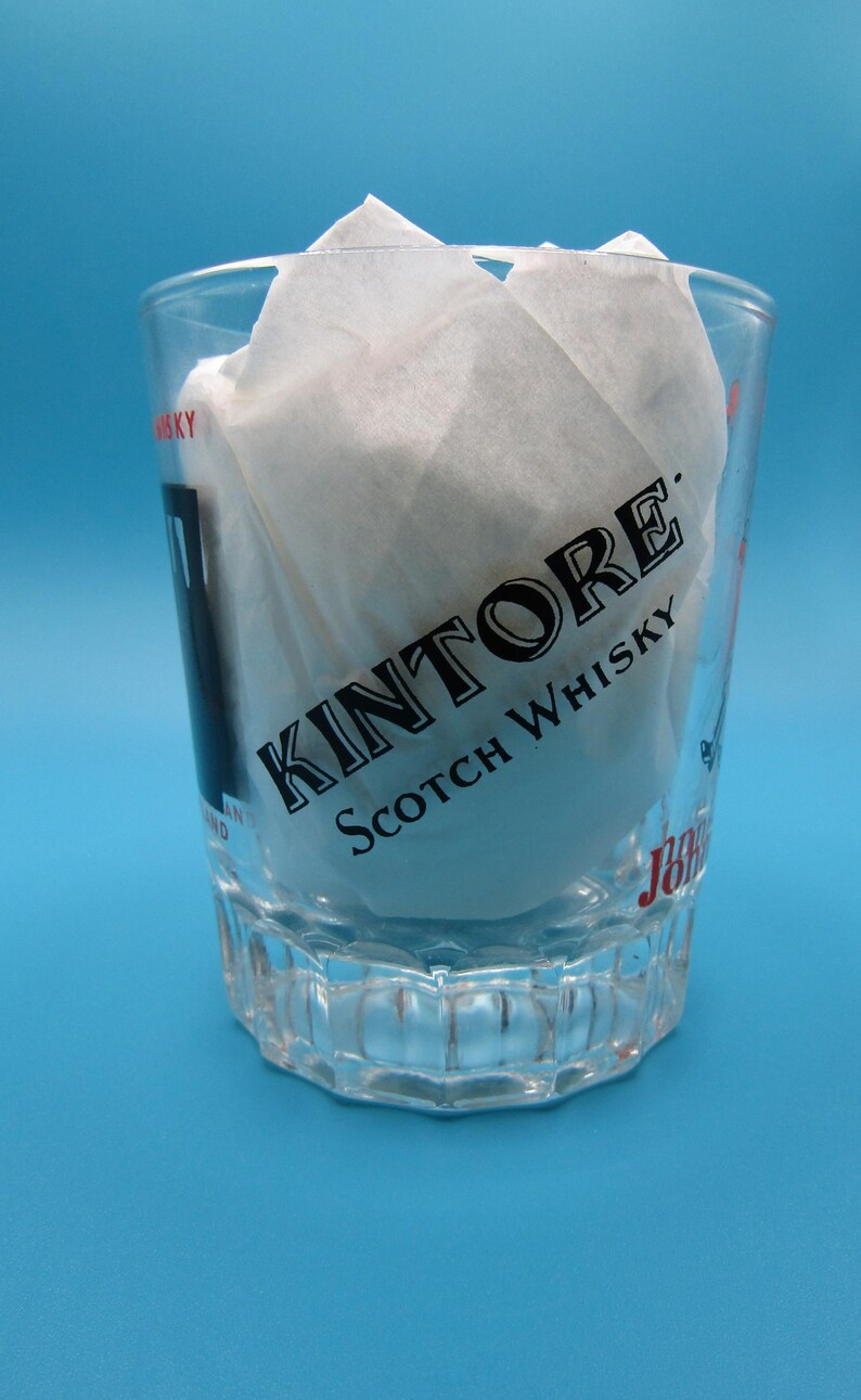Może przedstawiać: Przezroczysty kieliszek do w&oacute;dki z czarnym napisem "Kintore Scotch Whisky". Kieliszek jest wypełniony białą bibułą i stoi na niebieskim tle. Kieliszek ma wzorzystą podstawę.