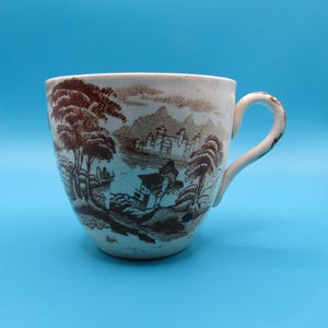 Puede incluir: Taza de té vintage con una escena de paisaje marrón impresa sobre un fondo blanco. La taza tiene un asa curva y un borde ligeramente acampanado. El paisaje presenta árboles, edificios y montañas.