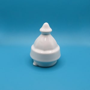Puede incluir: Una pequeña tapa de cerámica blanca brillante con una silueta suave, redondeada y escalonada. El objeto presenta una parte superior con un remate puntiagudo y una base circular ancha diseñada para encajar en un recipiente, con una superficie limpia.