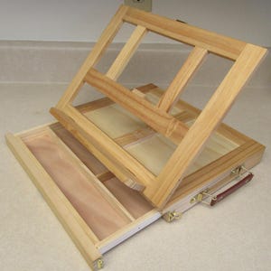 Könnte beinhalten: Ein offener hölzerner Künstlerstaffelei- und Aufbewahrungskasten. Die Staffelei ist aus hellem Holz gefertigt, mit einem Scharnierdeckel, der die Stütze der Staffelei bildet. Die Box hat Fächer für Kunstzubehör und einen Griff zum Tragen.