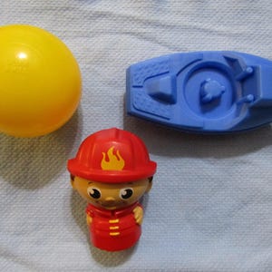 Può includere: Una palla di plastica gialla, un veicolo giocattolo blu e un giocattolo pompiere rosso con un'emblema a fiamma gialla. I giocattoli sono su una superficie bianca strutturata. Il giocattolo pompiere ha un casco e un'uniforme rossi.