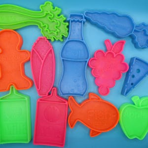 May include: A collection of colorful plastic cookie cutters in various shapes, including a gingerbread man, fish, apple, and a bottle labeled "Pop." Other shapes include a carrot, grapes, and a slice of pizza. The cutters are arranged on a blue surface.