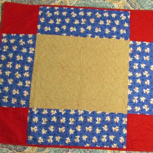 May include: A handmade quilt with a patchwork design. The quilt features a central tan square, surrounded by blue squares patterned with white shapes, and red borders. The quilt is likely made of cotton and has a quilted texture.