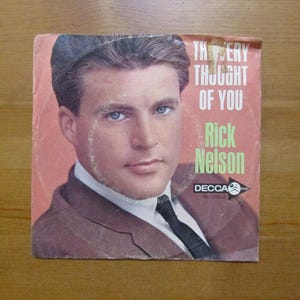 May include: A vintage square vinyl record sleeve featuring a portrait of Rick Nelson. The text reads The Very Thought Of You, Rick Nelson, and Decca No-31612. The background is a muted coral color, and the subject wears a brown suit and black tie.
