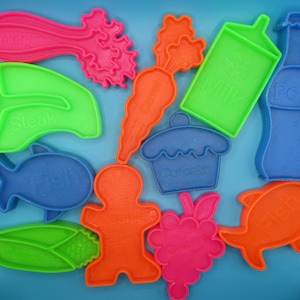May include: A collection of colorful plastic food-shaped cookie cutters. Includes shapes like a carrot, fish, celery, cupcake, milk carton, bottle, corn, gingerbread man, grapes, and steak. Each cutter is a different bright color, with text labels.
