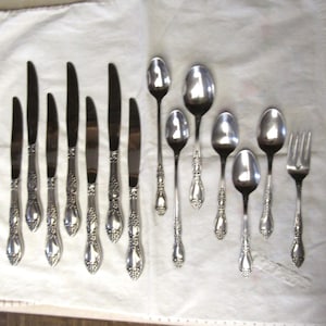 May include: A set of twelve pieces of silver-toned flatware featuring seven knives, four spoons of varying sizes, and one fork. Each handle is decorated with an embossed floral pattern at the base and along the stem, with polished metal surfaces.
