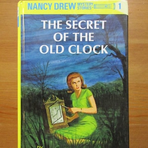 Puede incluir: Libro antiguo "El secreto del reloj antiguo" de Carolyn Keene, de la serie Nancy Drew. La portada muestra a una mujer con un vestido verde sosteniendo un reloj, con un fondo de bosque. El libro tiene un borde amarillo y un fondo azul.