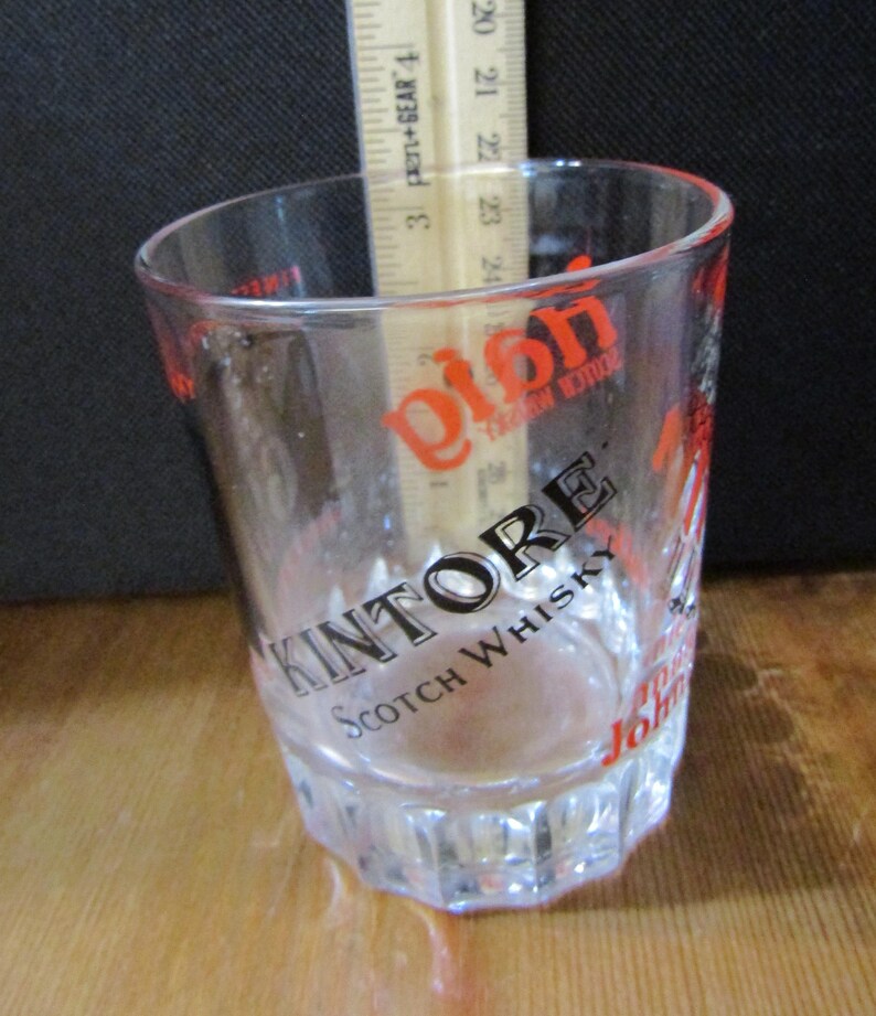 Może przedstawiać: Przezroczysty szklany kubek z napisem "KINTORE Scotch Whisky" w kolorze czarnym i czerwonym. Wewnątrz znajduje się drewniana linijka z podziałką w centymetrach. Szklanka ma fasetowaną podstawę.