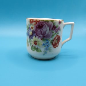 Op de afbeelding: Een witte theekop met een gouden rand en een oor. De kop is versierd met een bloemmotief met paarse rozen, rode en gele bloemen en blauwe accenten. De kop staat tegen een effen blauwe achtergrond.
