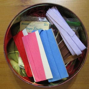 Op de afbeelding: Een ronde blikken doos met een rode rand, gevuld met naaibenodigdheden. Het bevat rode draden, gevouwen stoffen stroken in rood, roze, wit en blauw, en een pakketje "Rayon Elastic". Diverse andere naaiartikelen zijn ook zichtbaar.