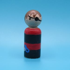 May include: A wooden peg doll in the shape of a pirate. The doll has a brown head with a painted eye patch and hair. The body is painted with red and black stripes, and a blue and white design.