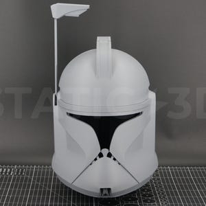 May include: A light gray, 3D-printed helmet, reminiscent of a clone trooper's helmet. The helmet features a rounded top, a visor, and a vertical antenna. The text "STATIC 3D" is visible on the background.