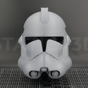 May include: A gray, 3D-printed Clone Trooper helmet, a piece of science fiction memorabilia. The helmet features a visor, cheek pieces, and a distinctive fin on top. The helmet is displayed on a grid surface.