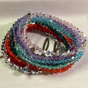 May include: A stack of colorful crystal bracelets with a silver charm. The bracelets are layered in a rainbow pattern, starting with clear, then light blue, teal, green, red, purple, and ending with clear again.