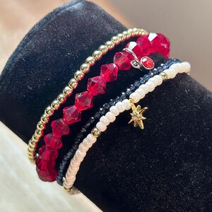 May include: A set of five bracelets featuring red, white, black, and gold beads. The bracelets are stacked on top of each other and include a small gold star charm.