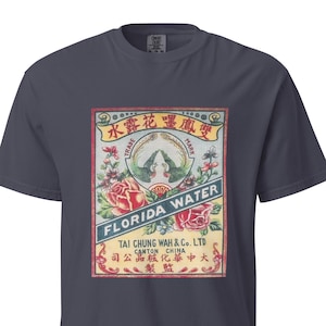 以下が含まれることがあります： ダークグレーの半袖Tシャツ。ヴィンテージのフロリダウォーターのラベルを模した長方形のグラフィックプリントが施されています。デザインには2羽の緑色の鳥、赤いバラ、そしてFlorida Water, Tai Chung Wah & Co. Ltd, Canton Chinaという文字が含まれています。