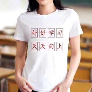 May include: A white t-shirt with red Chinese characters is worn by a person in a classroom. The text on the shirt is arranged in two rows of four characters each. The person is smiling.