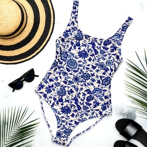 May include: A one-piece swimsuit with a blue floral pattern on a cream background. The swimsuit is paired with a straw hat with black stripes, black sunglasses, and black sandals. Palm leaves are also included.