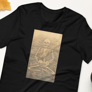 以下が含まれることがあります： 黒の半袖Tシャツ。中央には、様式化された波を背景に瞑想のポーズをとる人間の骨格が描かれた長方形のグラフィックプリントが施されています。骨格は細い線で描かれ、淡いベージュの色調で表現されています。