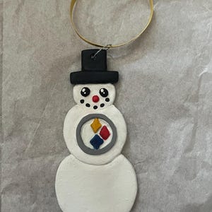 May include: A white snowman ornament with a black top hat and a Pittsburgh Steelers logo on its chest. The ornament has a gold hanger.