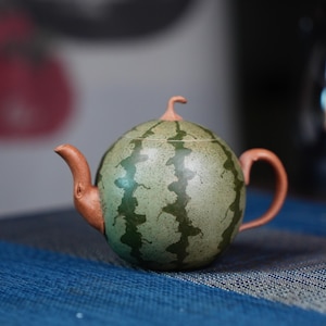 May include: A green and brown ceramic teapot shaped like a watermelon. The teapot has a spout and a handle.