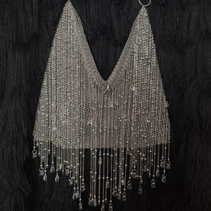May include: A silver-toned chainmail halter top featuring a deep V-neckline, constructed from fine interlocking metal links. The garment is adorned with numerous dangling silver chains and small, clear, teardrop-shaped beads along the bottom hem.
