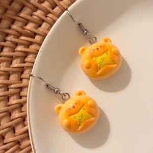 May include: A pair of orange bear-shaped earrings with yellow star-shaped centers. The earrings have silver-colored fish hook closures. The earrings are on a white plate.