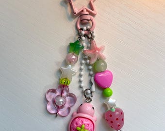 May include: A pink star-shaped lobster clasp keychain featuring three dangling strands of colorful beads. The charms include a translucent pink flower, a pink starfish, a pink heart, a small pink turtle with a strawberry shell, and a strawberry bead.